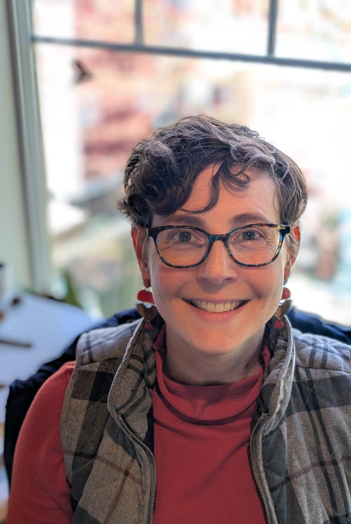 Alison Photo Alison sitting in front of a window with a blurry background. Alison is a white woman with short, curly brown hair and glasses. She is wearing a red orange turtle neck and glasses